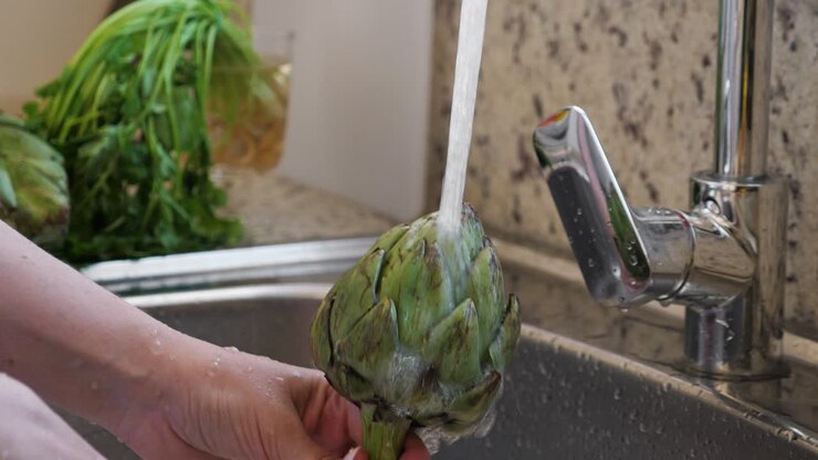 Woman washing artichokes in stainless steel sink with metal faucet. Cooking artichokes at kitchen