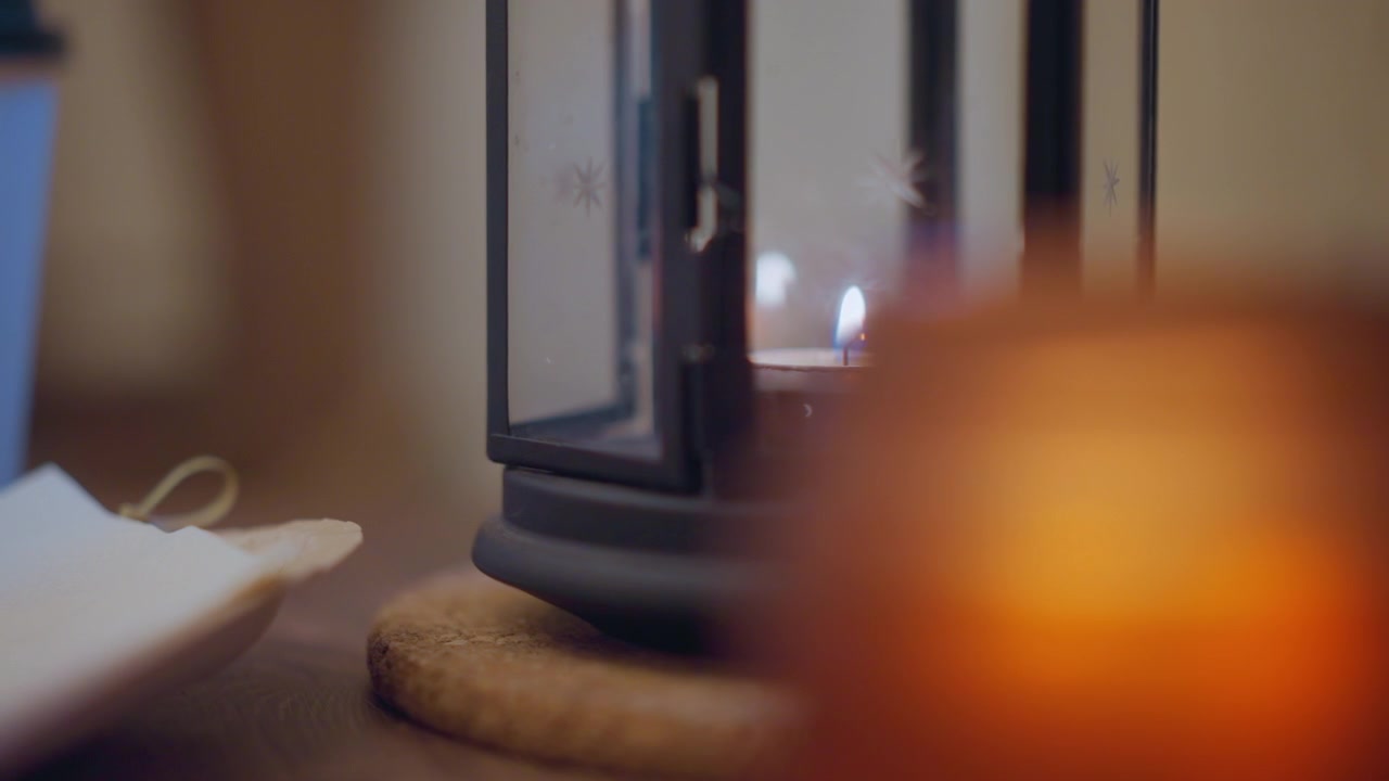Close up of lit candle in soft focus with ornate lantern and small white object on wooden surface. Candle flame flickers gently casting warm light through glass panels featuring snowflake