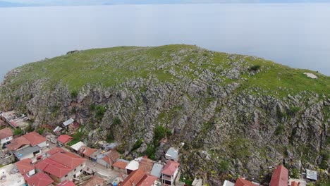 Drohnenansicht-In-Albanien,-Die-über-Eine-Kleine-Stadt-Mit-Häusern-Neben-Einem-Grünen-Hügel-Am-Ohridsee-Mit-Kristallblauem-Wasser-Fliegt