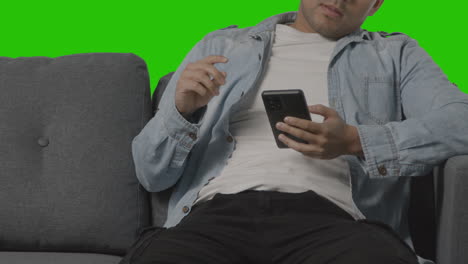 close up of bored young man sitting on sofa looking at mobile phone against green screen