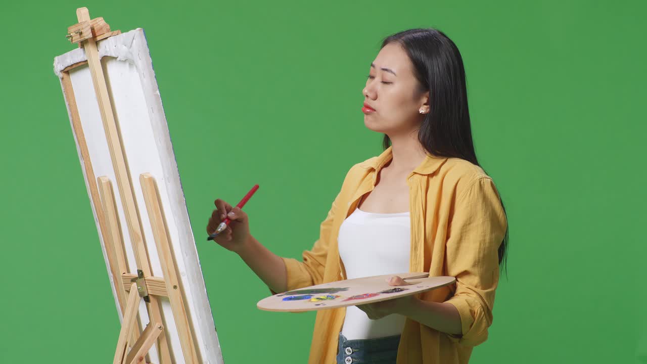 아시아 여성 예술가 는  을 들고 그림 개념 을 고민 하고 있으며, 그린 스크린 스튜디오 에서 오일 페인트 와 브러쉬 로 캔버스 에 그림 을 그리고 있다