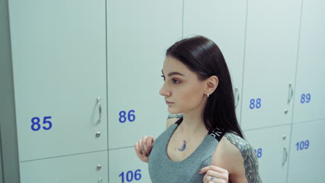 Attractive young woman looks at her slim body after workout in the dressing room. Dark-haired female with wing tattoos on her shoulders looks in the mirror in the sport center.