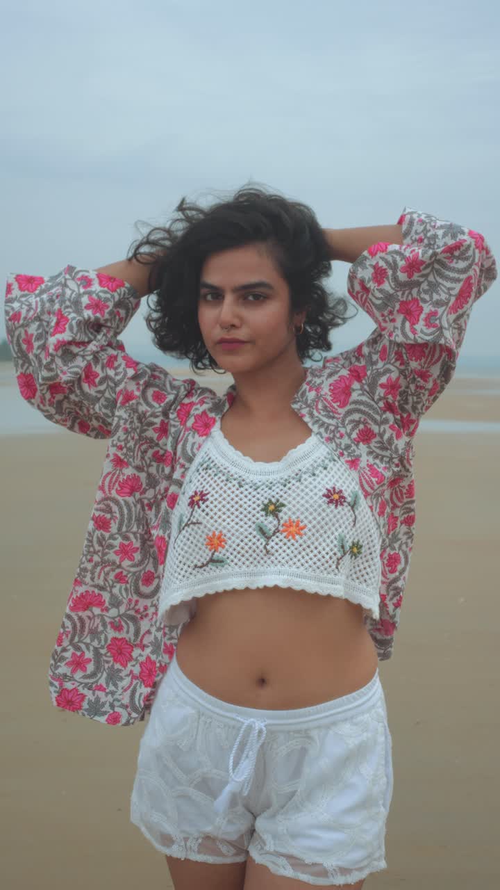 A young woman standing on a beach, wearing a floral shirt, embroidered crop top and white shorts, posing with her hands in her hair in a relaxed summer atmosphere