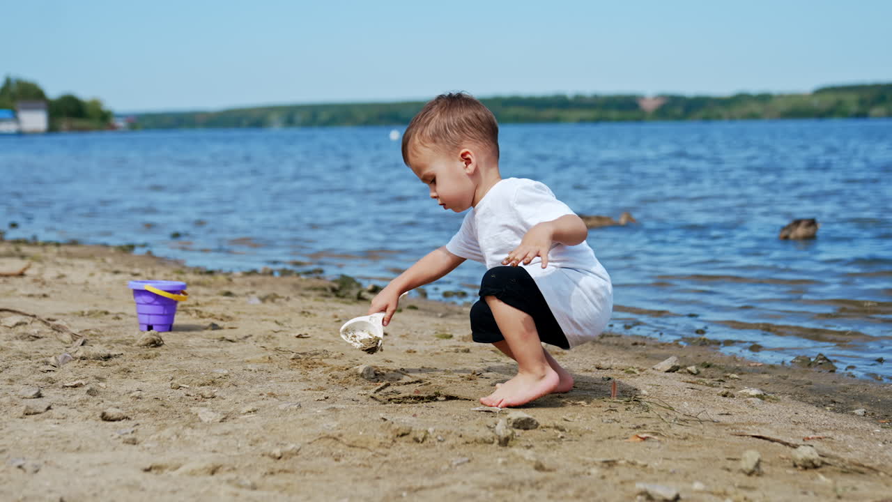 Little Caucasian boy in white t-shirt digging sand on the beach. Toddler working with shovel on the shore.