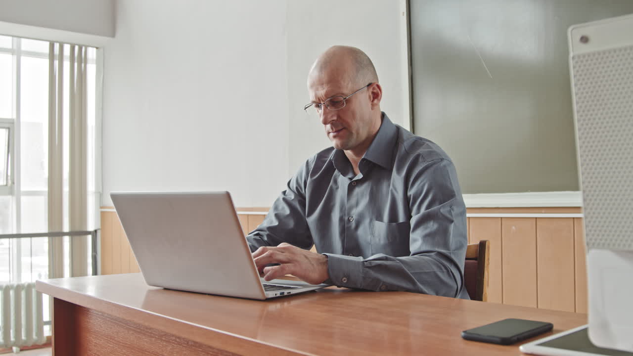 Male Mid-Adult Teacher Working on Laptop in Bright Classroom