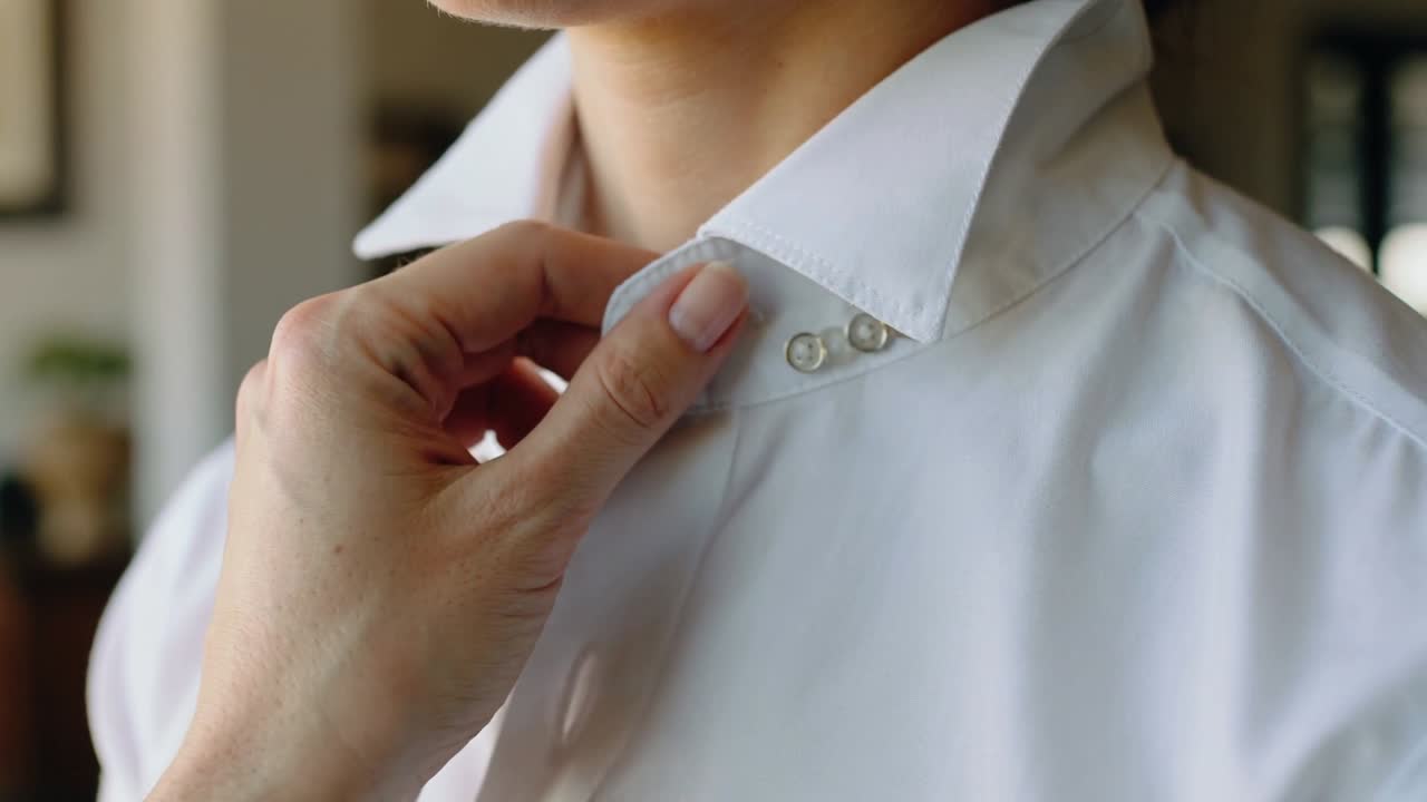 Mano con unghie rosa pallido che allaccia il colletto di una camicia bianca a casa, allineando i bottoni per fissare