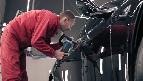 Hombre-Con-Traje-De-Trabajo-Rojo-Puliendo-Un-Coche-Negro-En-El-Centro-De-Automóviles