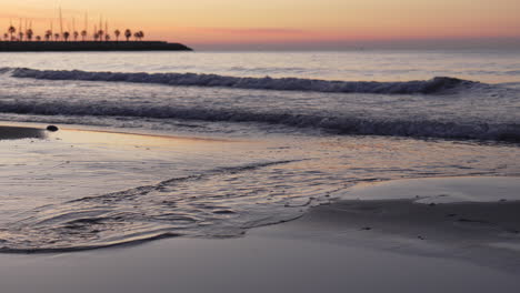 sunrise skyline is reflected in coastal waters that wash up onshore as a man walks across leaving footprints in sand