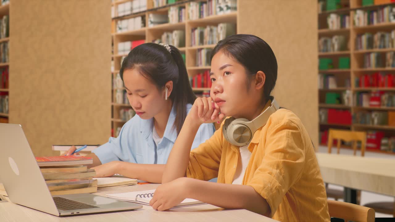 헤드폰을 착용한 아시아 여성 학생이 생각하고 주변을 둘러보며, 교실 친구와 함께 앉아서 노트북에 글을 쓰고, 도서관에 노트북을 가지고 있습니다.