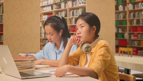 asian woman student with headphones thinking and looking around then raising her index finger while sitting with classmate writing into the notebook on table with a laptop in the library