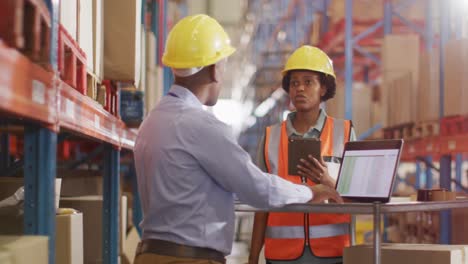 trabajadores afroamericanos masculinos y femeninos con casco y usando computadora portátil en el almacén