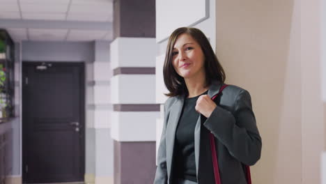 confident businesswoman in stylish suit with red bag standing in modern office hallway looking at camera with calm expression, casual professional appearance, sleek interior background, corporate vibe