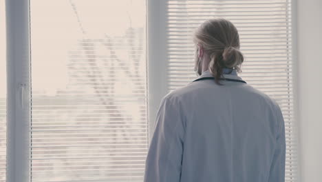 a young doctor with long, blond hair, looks worriedly out of the window and removes his mask