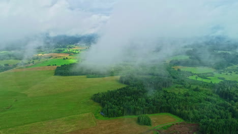 Misty-green-fields-and-forests-under-a-cloudy-sky,-tranquil-rural-landscape,-aerial-view