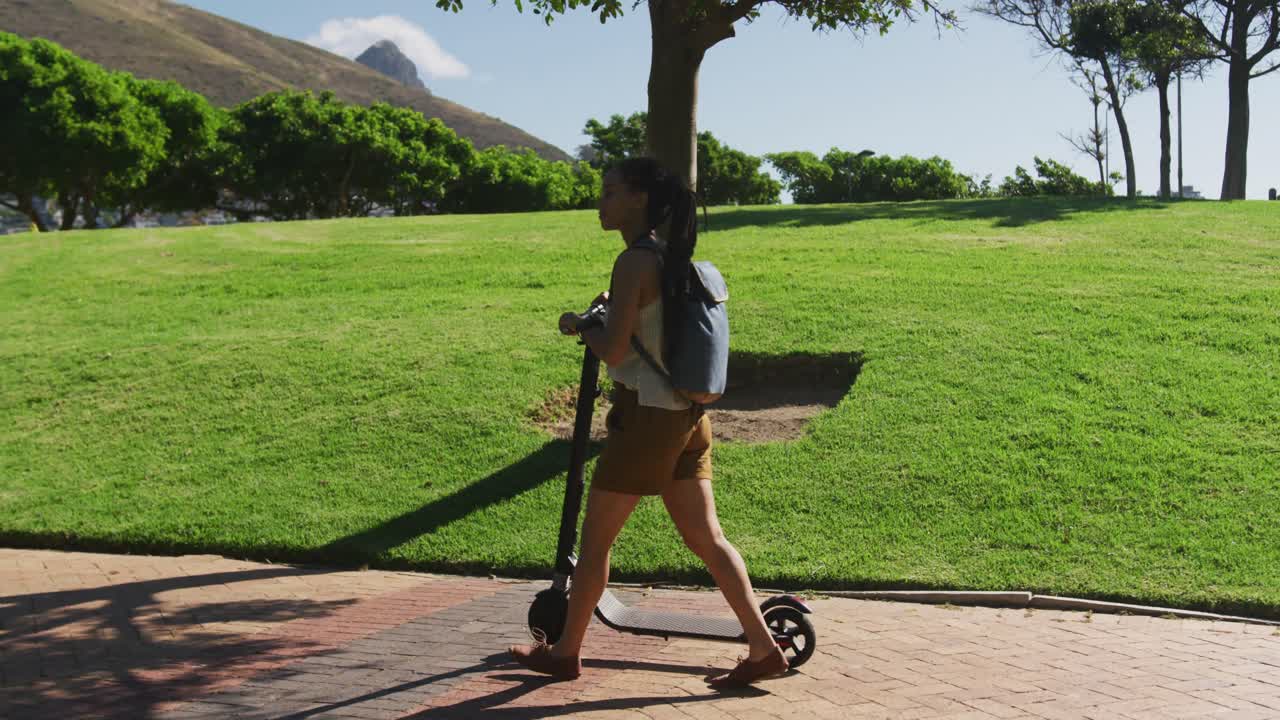 Mujer afroamericana con mochila montando en patinete en un parque.