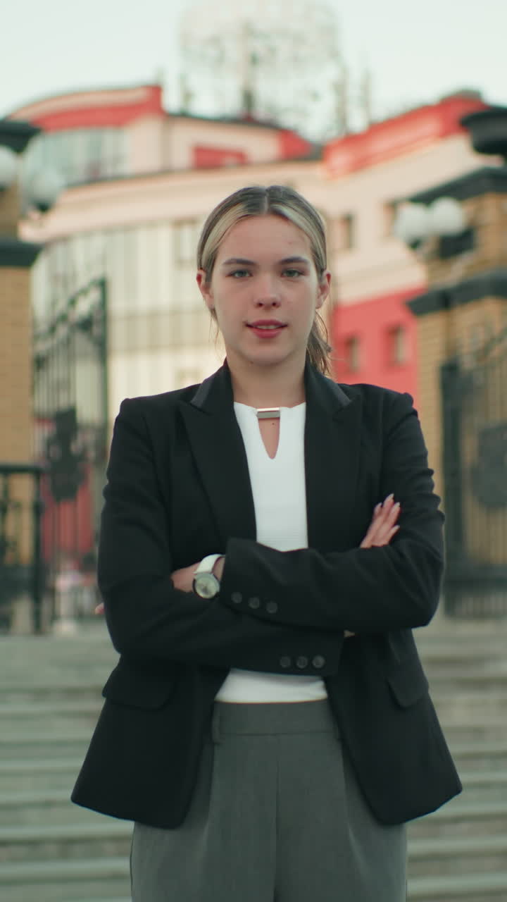 Young woman standing confidently on steps in busy urban environment, arms crossed, dressed in professional attire, exuding elegance and determination, with background of buildings
