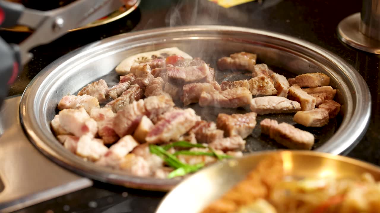 Gloved hand cuts sizzling pork belly on tabletop grill, bright lighting, shallow depth of field