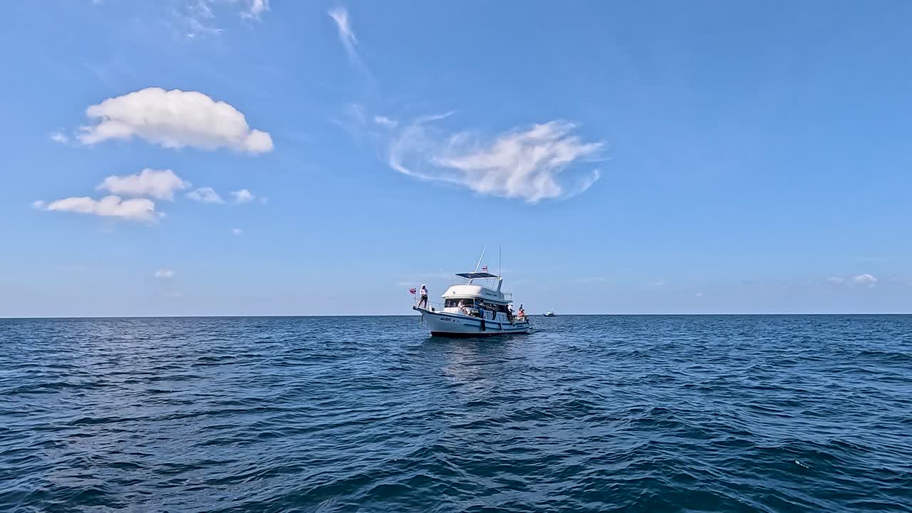 A fishing boat with a person aboard moves steadily toward the camera across calm ocean waters under bright daylight and clear blue sky