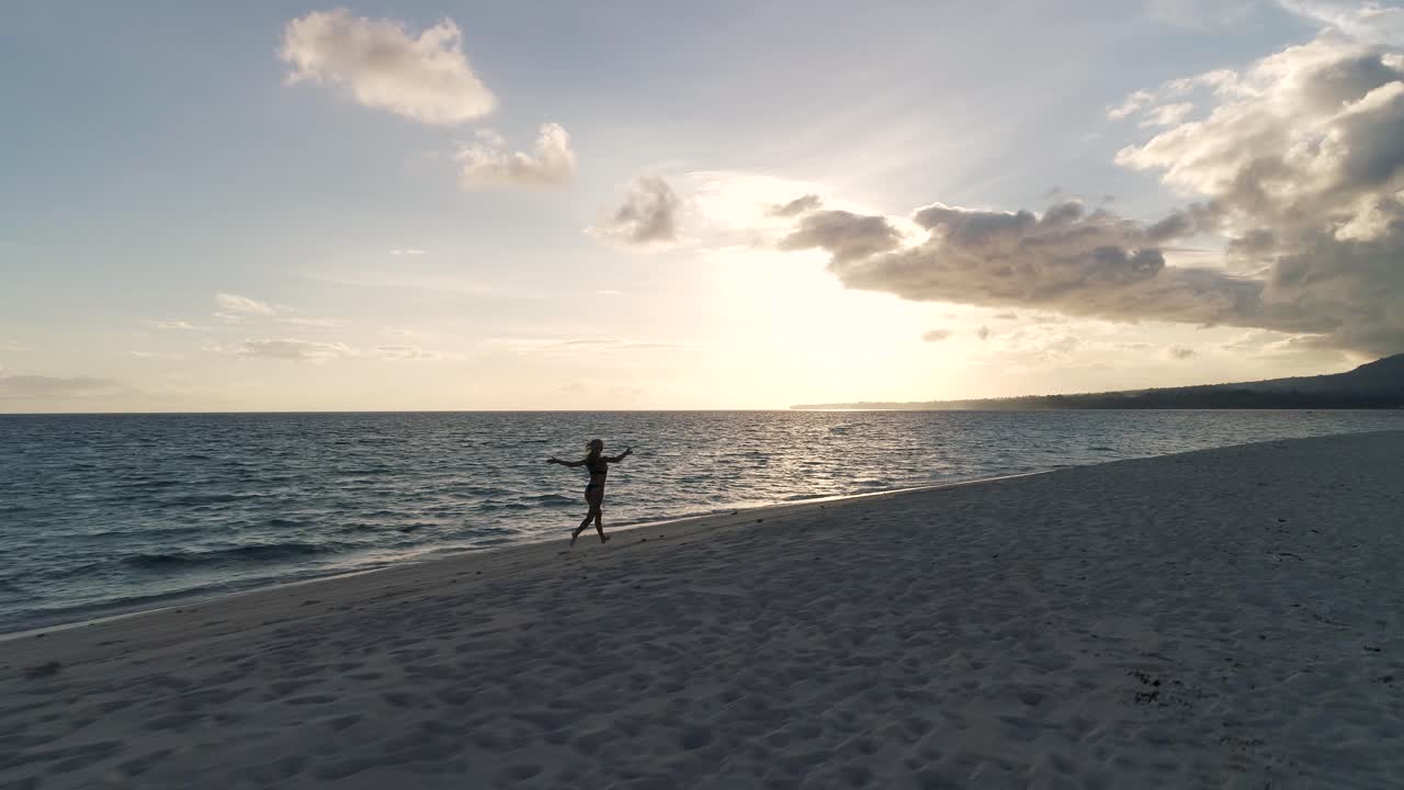 Carefree woman enjoys running on tropical beach at dawn, spreading arms, aerial
