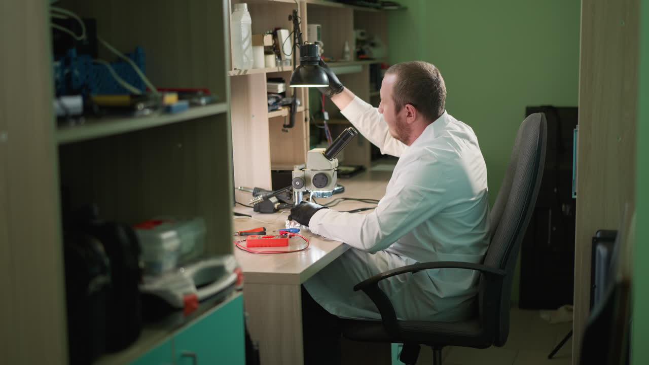 una vista cercana de un técnico en una bata de laboratorio blanca y guantes de mano negros ajustando una lámpara de escritorio y continúa el circuito de trabajo bajo un microscopio usando un pequeño picker de hierro con un estante al lado