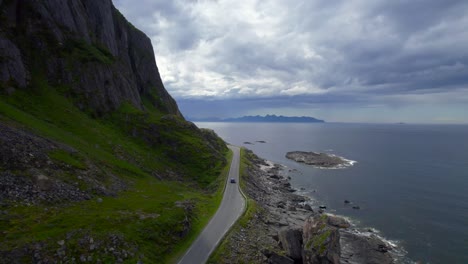 Low-Flyover-shot-of-a-person-hiking-and-revealing-the-scenic-route-on-Andøya,-with-a-vehicle-driving-along-the-curvy-rocky-coastline