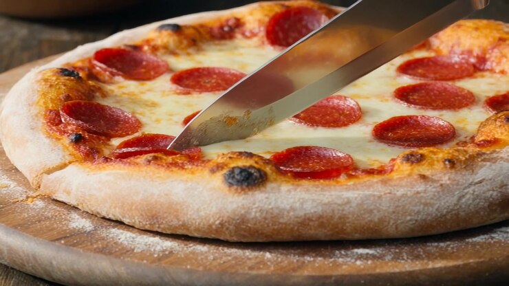 Mano che affetta con un coltello da chef, toccando la crosta, tagliando una pizza ai peperoni per servirla su un tagliere di legno.