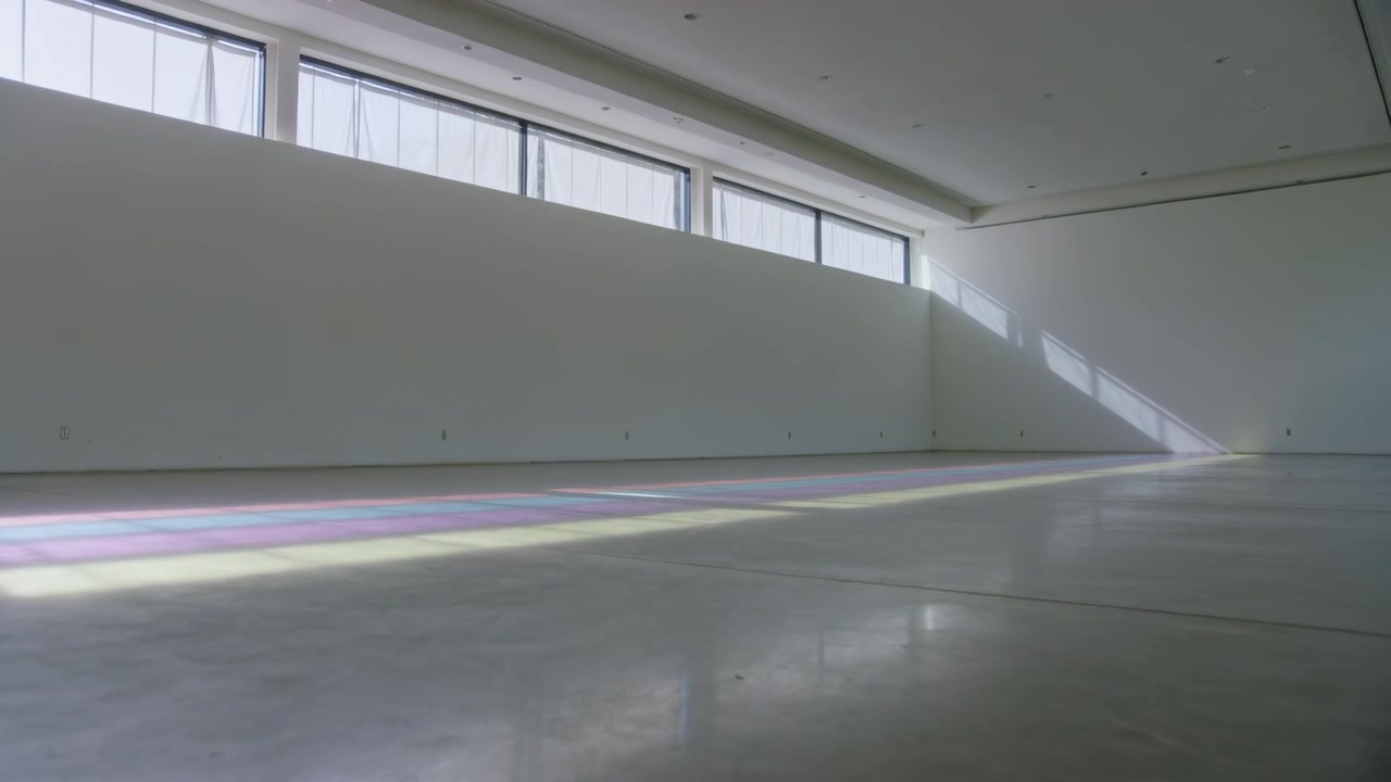 Faixa colorida em movimento atravessando o chão de concreto em uma galeria, com o sol passando por janelas altas.