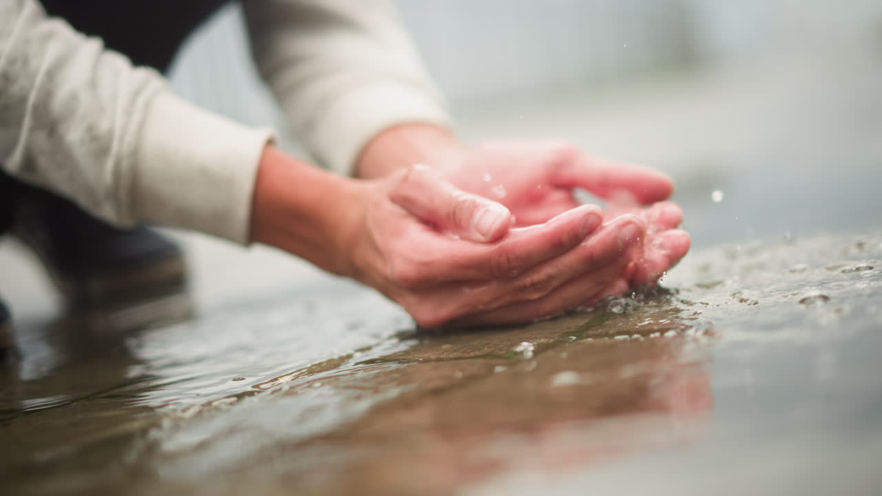 백인 손이 빗물 웅덩이 포장 도로를 만지며 부드럽게 물을 뜨고, 흐릿하게 보이는 흐린 하늘, 부드러운 스웨터 소매, 반사되는 표면, 명상적인 고독, 촉각적인 탐구, 도시의 아침 분위기