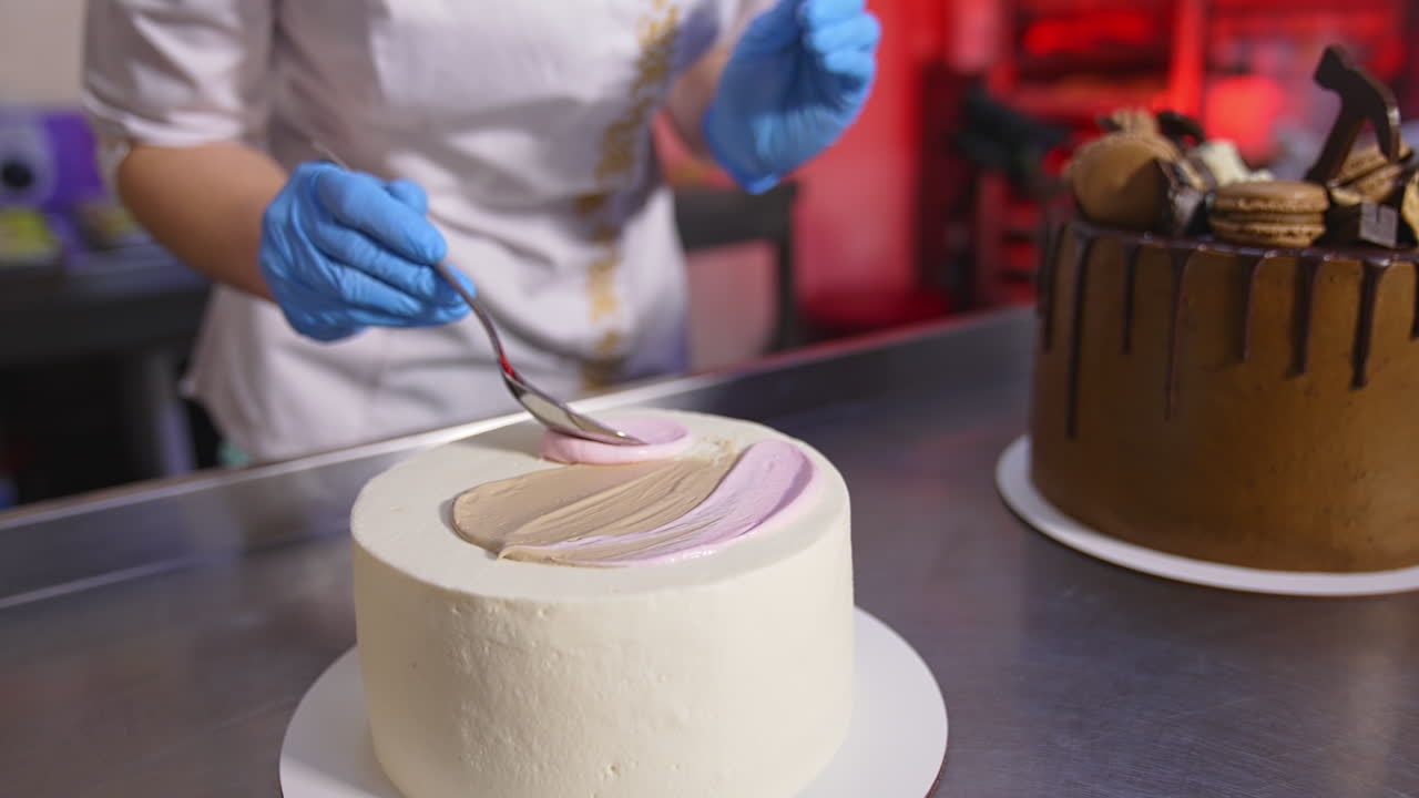 Pastry cook applies white cream with a spoon on top of the white cake. Close up. Chocolate cake at the table at backdrop.
