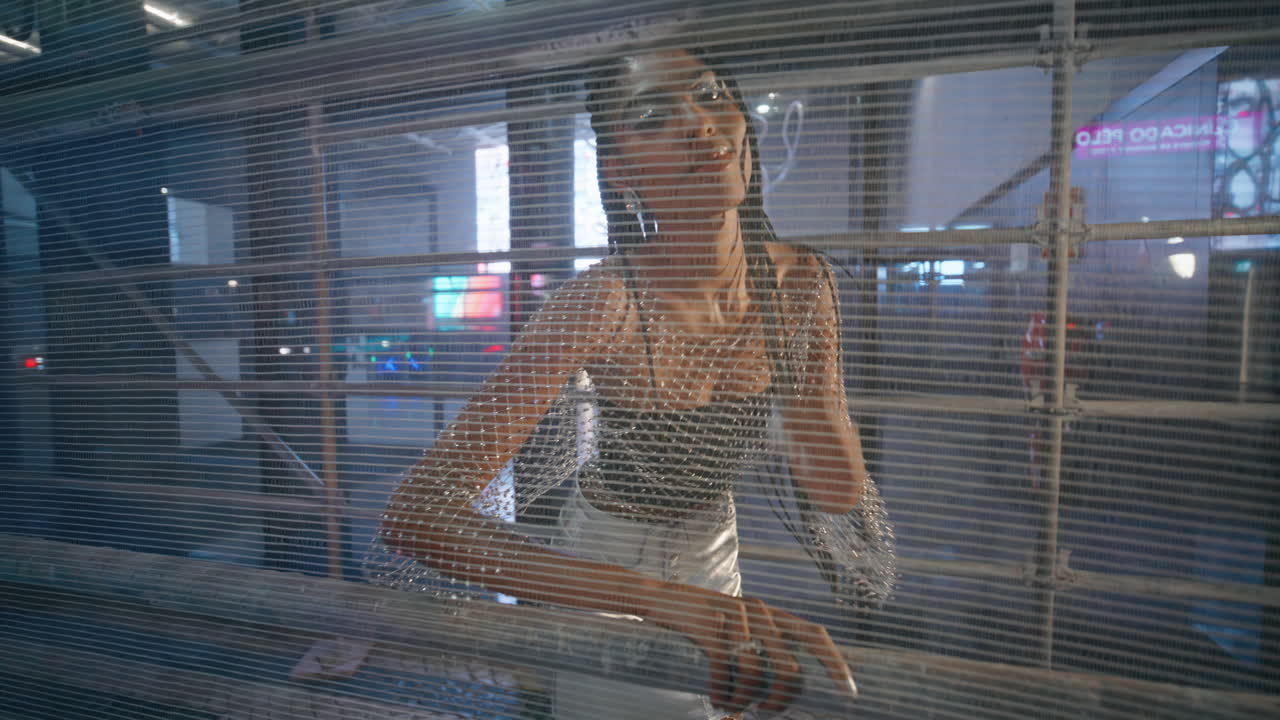 자신감 있는 여성 은 느 한 자세 로 밤 도시 를 포즈 하고 있는 금속 격자 를 보고 있다