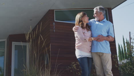 Portrait-Of-Smiling-Senior-Couple-Standing-Outdoors-In-Front-Of-Dream-Home-House-With-Garden