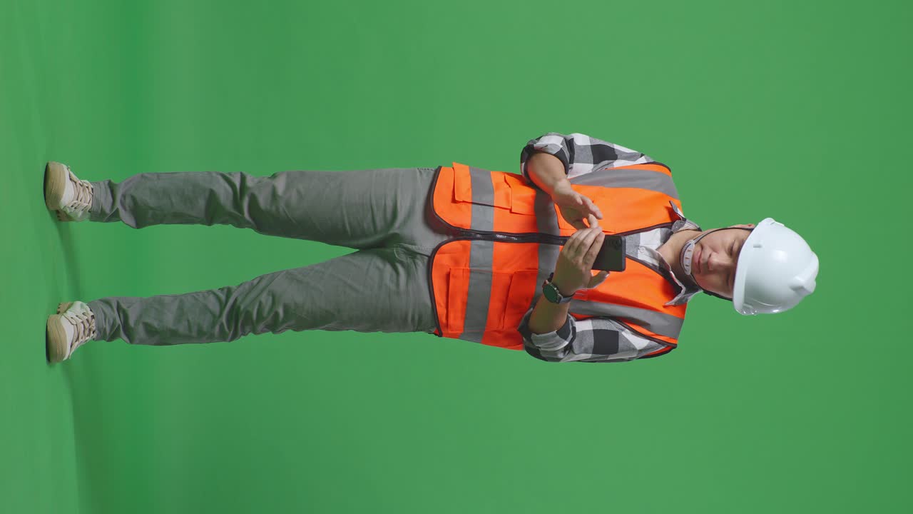 Full Body Of Asian Male Engineer With Safety Helmet Using Smartphone While Standing In The Green Screen Background Studio