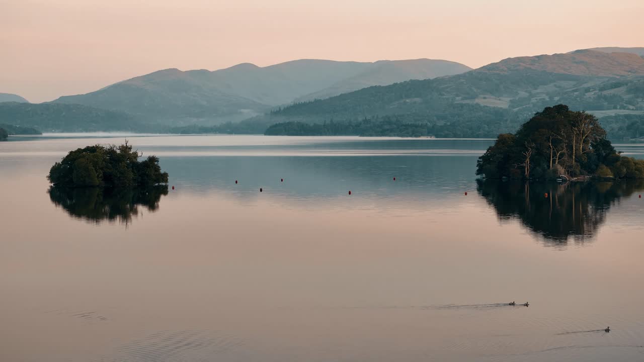 Aerial dolly of ducks swimming on Windermere Lake, Lake District England as rolling hills float down to water and reflect with orange sunset sky