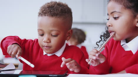 Un-Niño-Y-Una-Niña-Usando-Una-Tableta-Y-Un-Lápiz-óptico-En-Un-Escritorio-En-Un-Aula-De-Escuela-Infantil,-De-Cerca