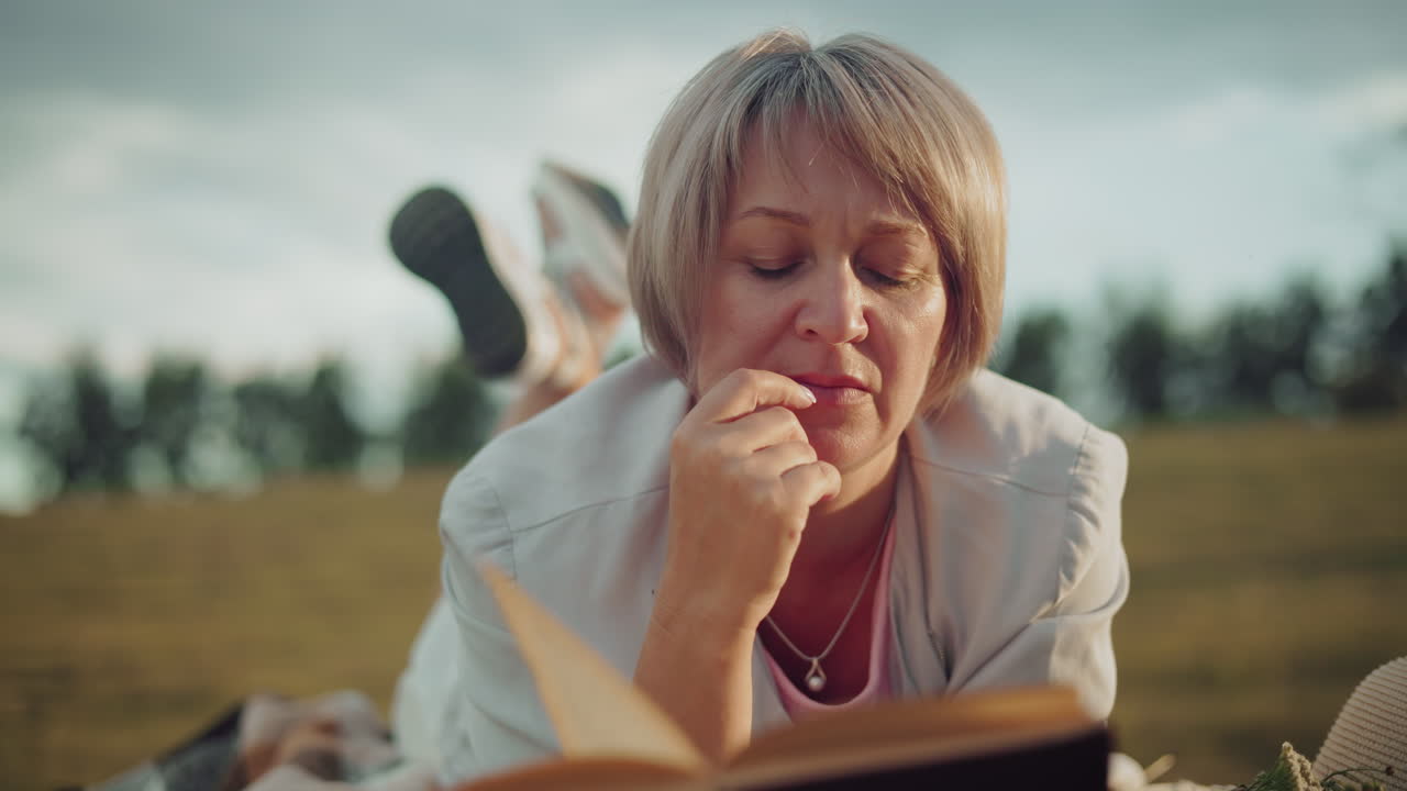 Close-up of woman thoughtfully reading book with hand on chin, resting on hay bale in vast open field, surrounded by nature and distant trees