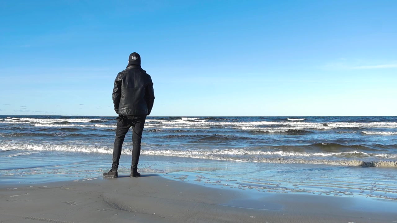 Mann med svarte klær og skinnjakke ser på bølger i Østersjøen som beveger seg i sakte film på en solrik dag på Vääna strand, tenker og går sakte bort. Reflekterende vann, sandete.