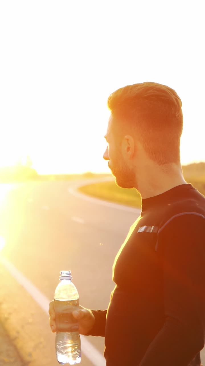 Athletic man drinking water during sunset while standing on a road with bright sunlight