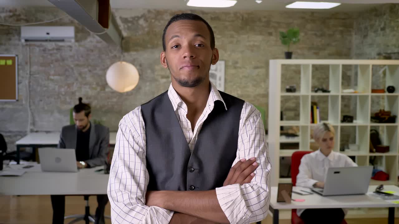 Young african businessman is standing with arms crossed and watching at camera in office, thinking concept, colleagues are working with laptops, work concept