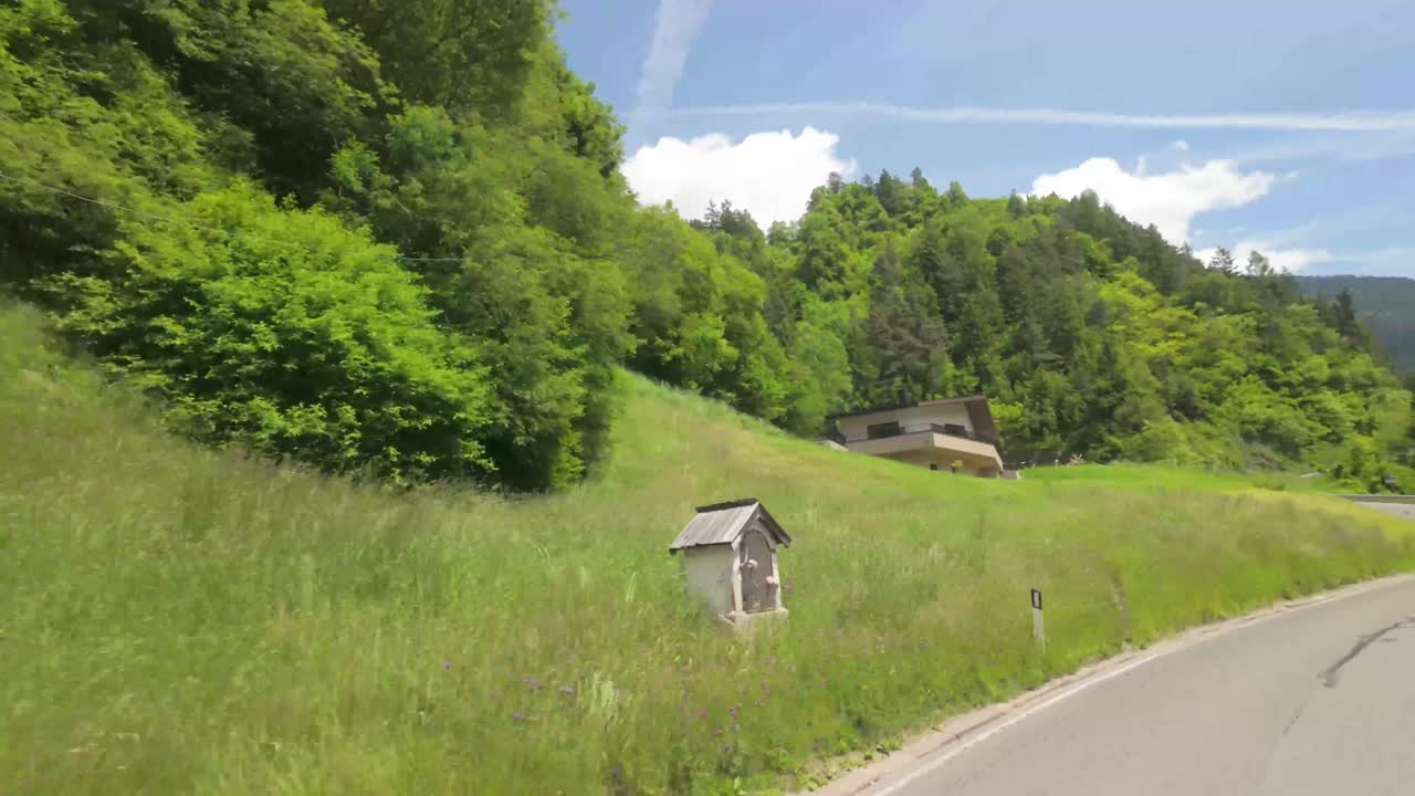Tracking shot of campervan driving through a picturesque alpine village in Italy, passing traditional gasthaus and rustic buildings nestled in mountain scenery.
