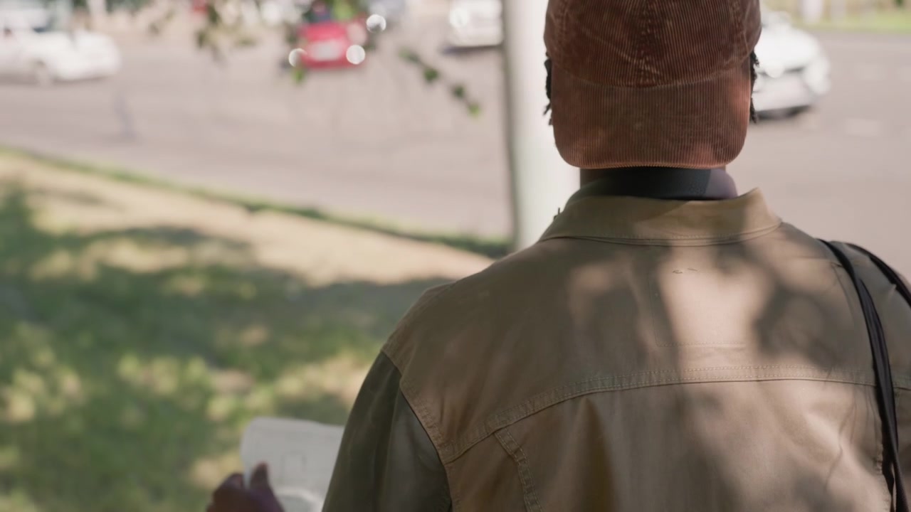 Close up of man holding white paper outdoors with green grass and parked cars in background. Man wears brown jacket cap carries bag while examining document under natural daylight. Soft