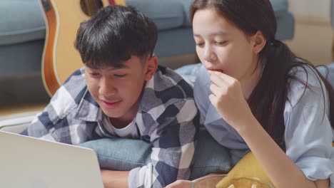 close up of asian teen couple using a laptop while lying on carpet on the floor at home. eating snacks watching movie, speaking, and enjoying time together