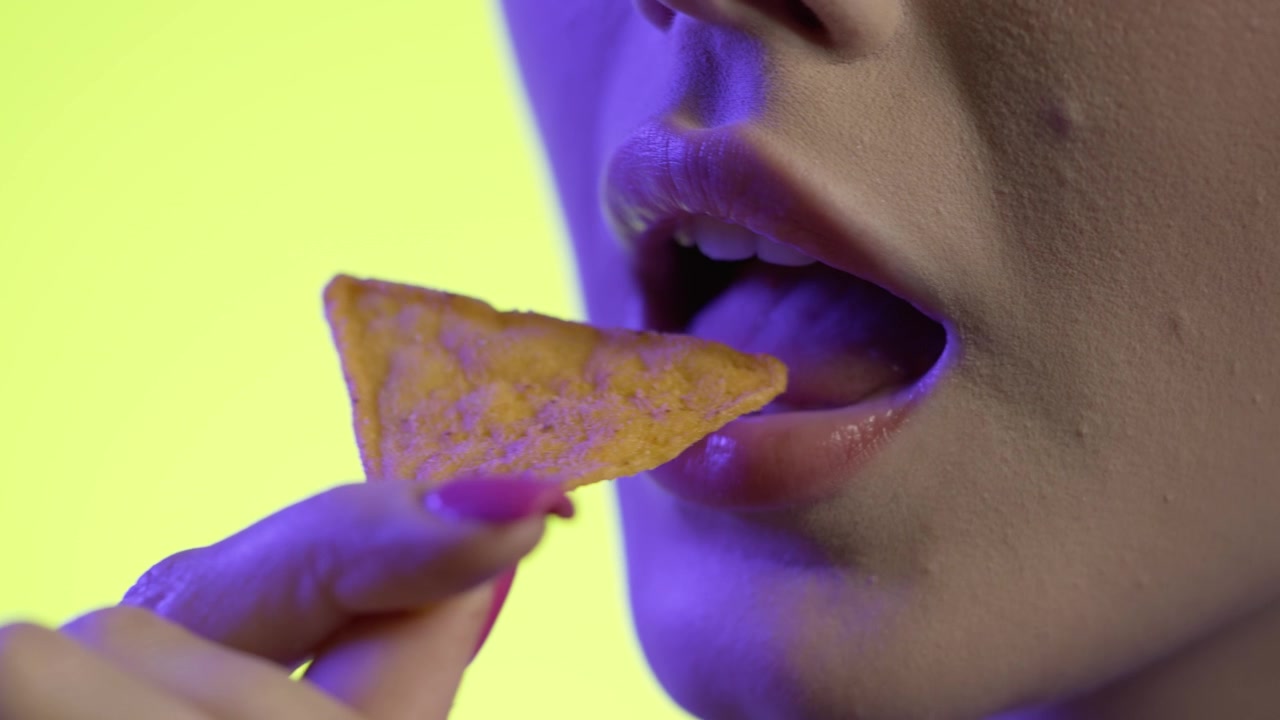 mulher jovem comendo um chip de tortilla, mastigando a boca em close-up em fundo amarelo, iluminação rosa