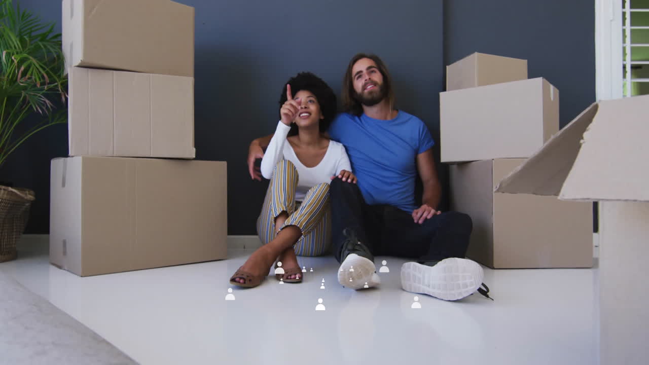 Sitting on floor, couple surrounded by moving boxes, smiling and pointing