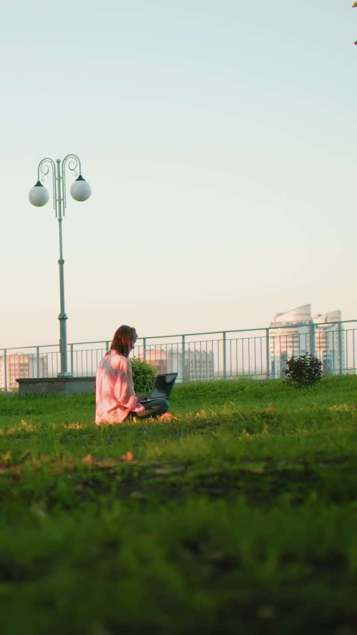 잔디에 앉아 노트북으로 일하는 여성, 배경에는 조명 기둥, 철로, 도시 건물, 도시 요소와 자연을 결합하는 평화로운 설정