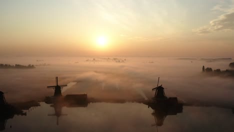 the drone is flying away from the windmills of the zaanse schans during sunrise with lots of mist and the silhouettes of the windmills inthe netherlands aerial footage 4k