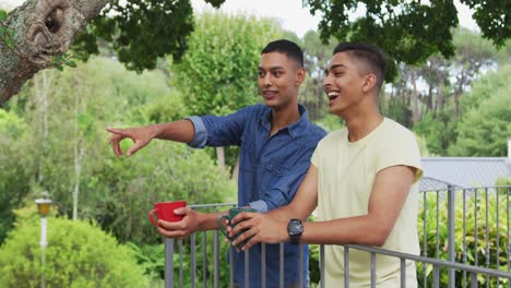 Mixed-race-gay-male-couple-standing-in-garden-drinking-cups-of-coffee-talking-and-laughing