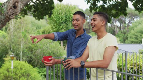 mixed race gay male couple standing in garden drinking cups of coffee talking and laughing