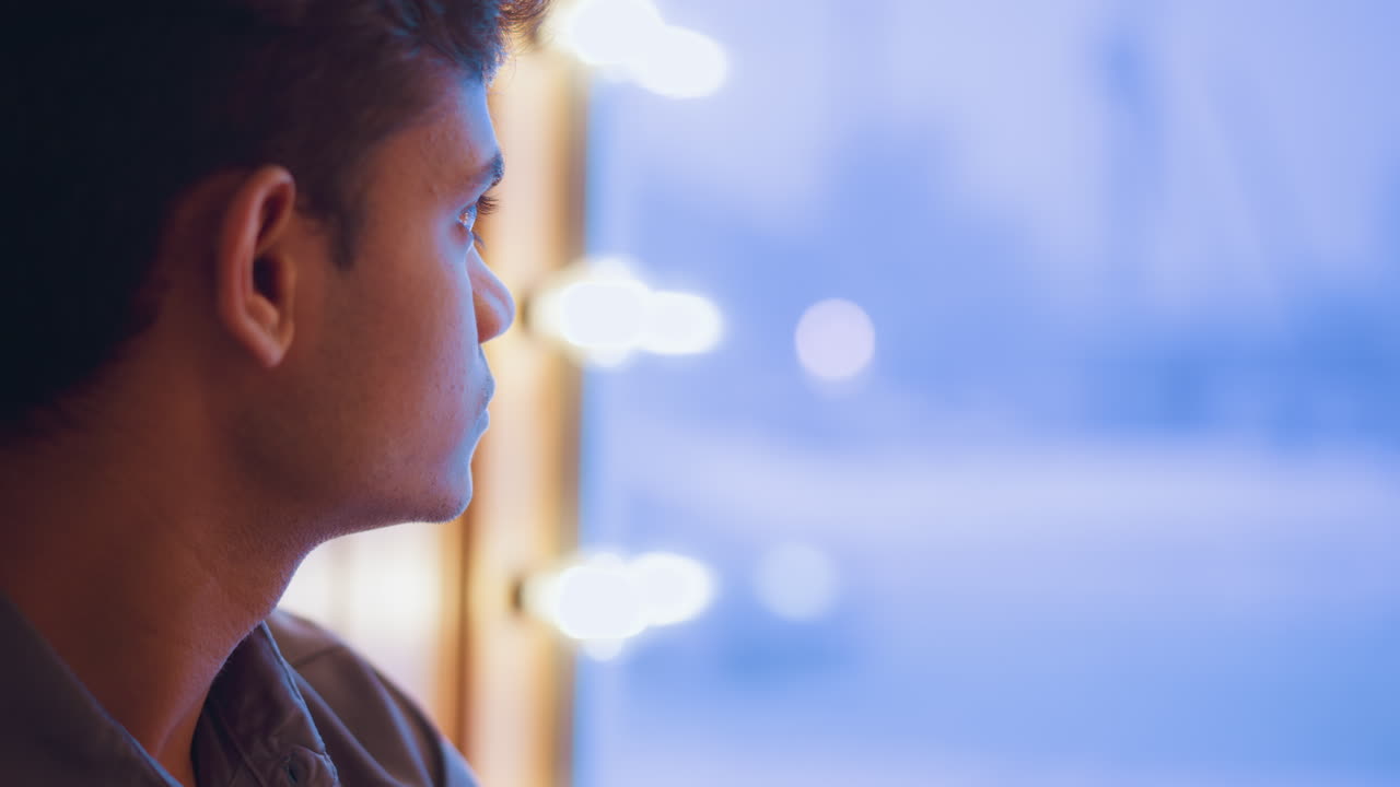 Young man seen from behind looking through window bathed in soft blue light, appearing lost in thought or calm observation, capturing tranquil indoor mood of solitude, stillness, and quiet reflection