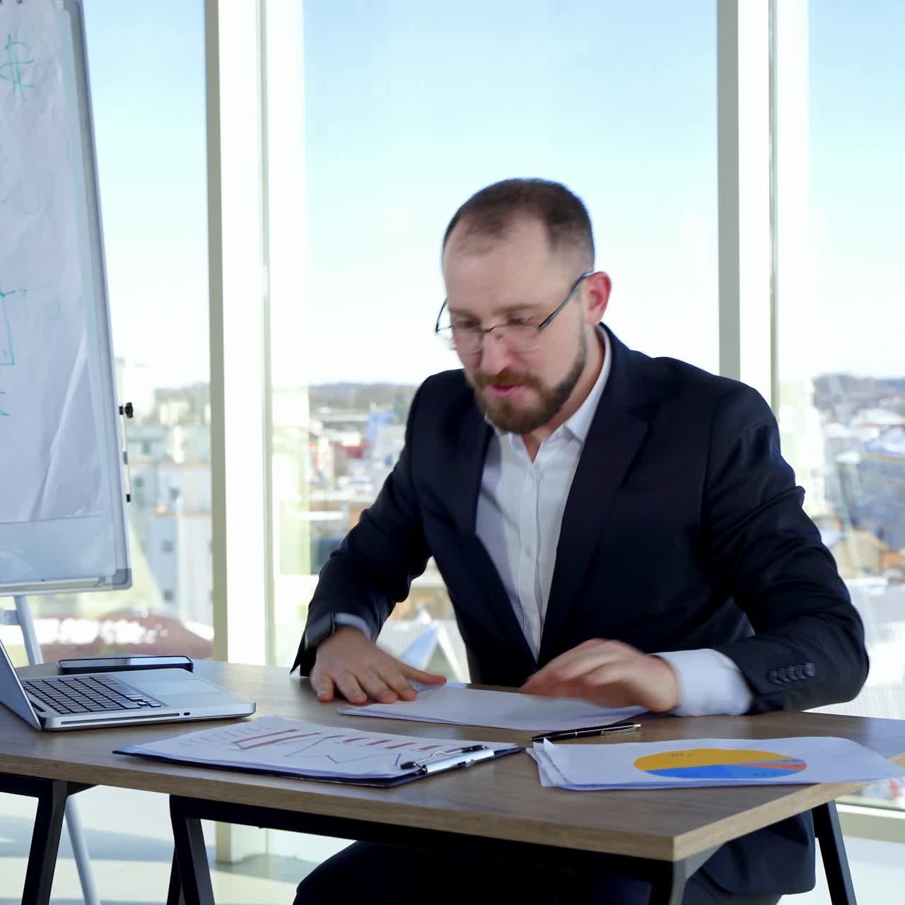 Nervous businessman in office. Irritated man entrepreneur sitting at table and tearing papers throwing them to the sides on the background of window