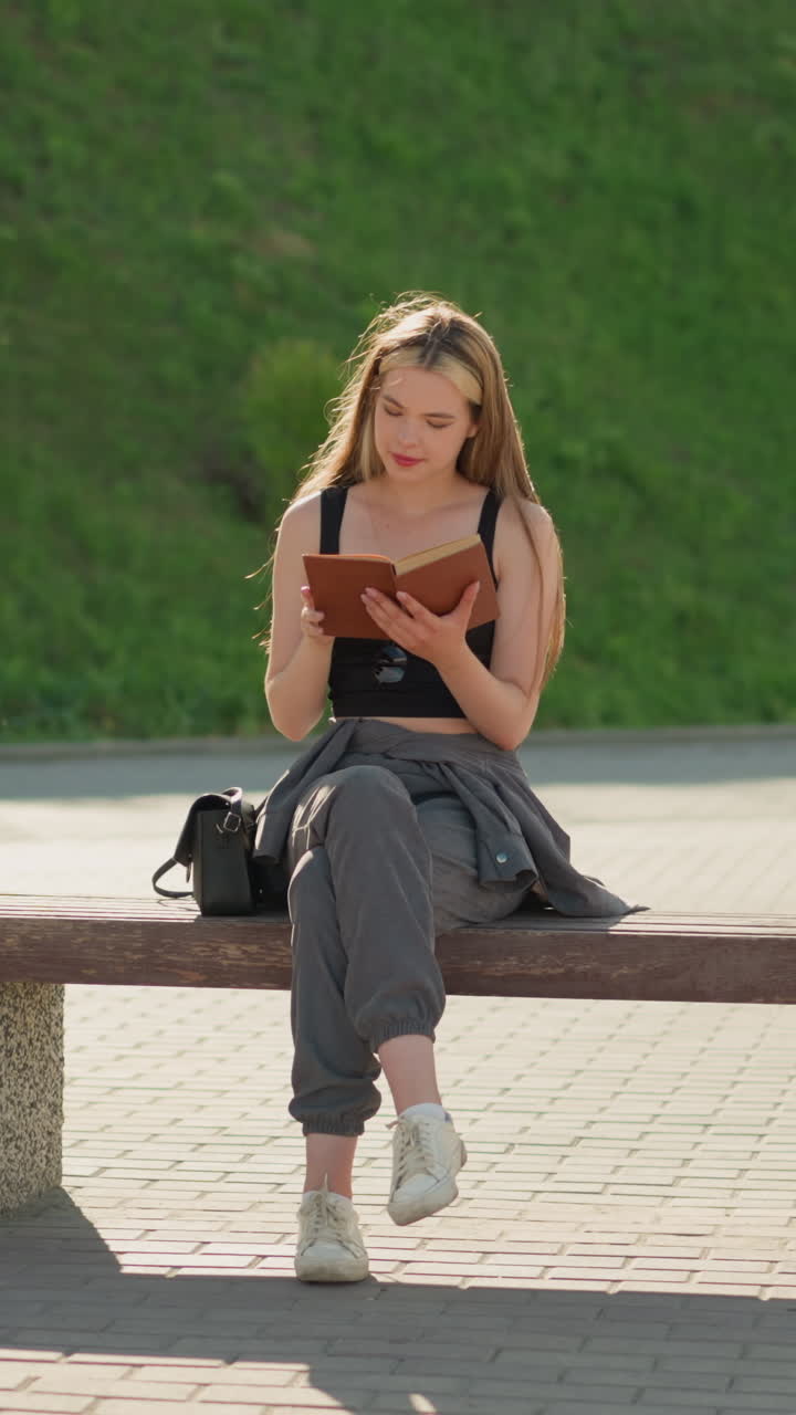 Woman seated outdoors on a bench with a smile, reading a book with legs crossed, she holds the book gently as the sun illuminates her, the background features two distant people, a light pole
